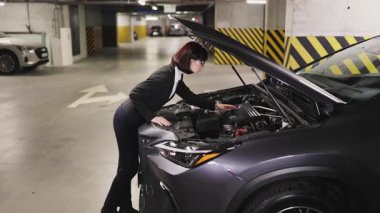 Beyaz iş kadını yeraltı otoparkındaki araba motorunu inceliyor. İş elbiseli genç bir kadın araç tamirine odaklanıyor..