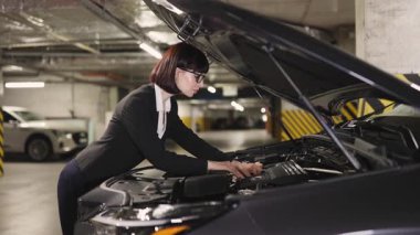 Resmi giyimli beyaz iş kadını yeraltı otoparkındaki araba motorunu tamir ediyor. Tamire odaklanmış, gözlük takıyor ve kaputun yanında duruyor, elleri motorda görünüyor..