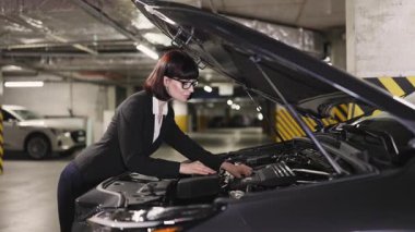 Beyaz kadın iş kadını yeraltı otoparkındaki araba motorunu inceliyor. Araç tamiri üzerinde çalışırken odaklanmış görünüyor. Bağımsızlık kavramı, problem çözme ve otomotiv bakımı.