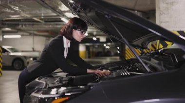 30 'lu yaşlardaki kendine güvenen beyaz iş kadını yeraltı garajında kaputun altındaki araba motorunu inceliyor. Profesyonel kıyafet ve odaklanmış ifade özgüven ve kararlılığı ifade eder..