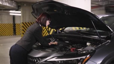 Beyaz kadın iş kadını yeraltı otoparkındaki araba motorunu inceliyor. Araç tamiri üzerinde çalışırken odaklanmış görünüyor. Bağımsızlık kavramı, problem çözme ve otomotiv bakımı.