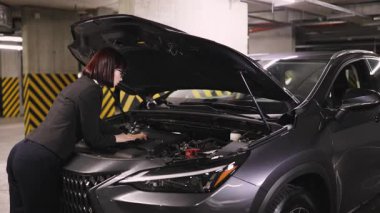 Beyaz iş kadını yeraltı otoparkındaki araba motorunu tamir ediyor, bağımsızlığını ve yetkilerini gösteriyor. Görüntü, profesyonel giyimli, mekanik sorun çözen modern kadını yakalıyor..
