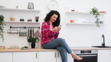Sıradan kıyafetler giyen Afro-Amerikalı kadın mutfak tezgahında oturmuş elinde akıllı telefonuyla gülümsüyor ve keyfine bakıyor..