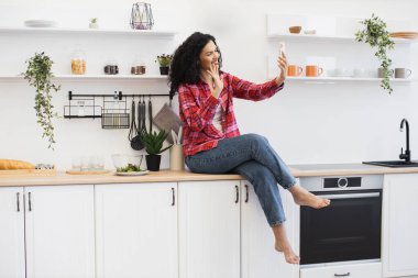 Mutfak tezgahında oturan Afrikalı kadın, günlük kıyafet. Gülümseme, el sallama, akıllı telefonu görüntülü konuşma için kullanma, aydınlık ev ortamında dostane atmosfer, modern dekor, taze yeşillik..