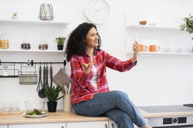Gülümseyen Afrikalı kadın mutfak tezgahında oturmuş elinde akıllı bir telefonla selfie çekiyor, mutluluk ve rahatlama ifade ediyor, mutfak gereçleri, bitkiler ve sağlıklı bir salata ile çevrili..