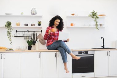 Kıvırcık saçlı Afrikalı kadın tezgahın üzerinde oturuyor, modern parlak mutfakta, dekoratif eşyalarla çevrili bir şekilde dizüstü bilgisayar kullanıyor, sıcak bir ortam yaratıyor..