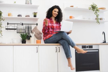 Gündelik giysiler içinde, bilgisayarı mutfakta kullanan, internetten iletişim kuran, birden fazla iş yapan Afrikalı Amerikalı bir kadın.