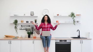 Modern mutfakta oturan Afrikalı kadın dizüstü bilgisayar kullanıyor, meditasyon yapıyor, sakin bir atmosfer yaratıyor. Beyaz minimalist mutfak tarzı, kapalı alan bitkileri dekore eder.