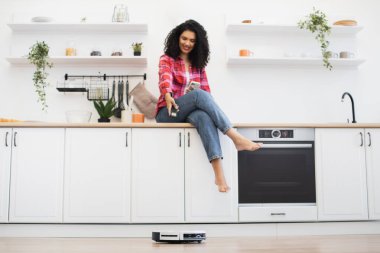 Mutfak tezgahında oturan Afrikalı kadın robot elektrikli süpürge kullanıyor, mutlu ve rahat görünüyor. Modern aletlerle aydınlık, temiz mutfak ortamı elverişli.