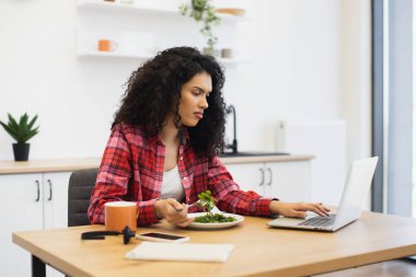 Genç Afrikalı Amerikalı kadın ev mutfağında dizüstü bilgisayarla çalışırken sağlıklı yemek yiyor..