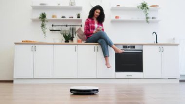 Modern mutfakta oturan Afrikalı kadın gülümseyerek robot süpürgeyi kontrol ederken akıllı telefon kullanarak teknolojik kolaylığı ve ev verimliliğini destekliyordu.