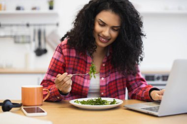 Afro-Amerikalı genç bir kadın mutfakta salata yiyor, laptopla rahatlamış bir şekilde çalışıyor. Sağlıklı yaşam tarzına, üretkenliğe ve dengeli diyete önem veren günlük ortam.