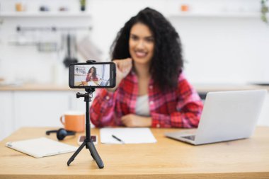 Genç Afrikalı kadın akıllı telefon, dizüstü bilgisayar ve kırtasiye malzemeleriyle mutfak ortamında içeriği kaydediyor. Evde teknolojiyi kullanarak sosyal medya içeriği oluşturur.