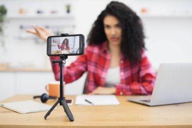 Afrikalı bir kadın gündelik giysiler içinde oturmuş akıllı telefon tripodu kullanarak video kaydediyor..
