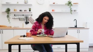 20 'li yaşlarda Afro-Amerikalı bir kadın açık mutfakta sağlıklı yemek yerken bilgisayarla çalışıyor..