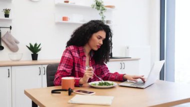 Afro-Amerikalı genç bir kadın mutfakta salata yiyor, laptopla rahatlamış bir şekilde çalışıyor. Sağlıklı yaşam tarzına, üretkenliğe ve dengeli diyete önem veren günlük ortam.