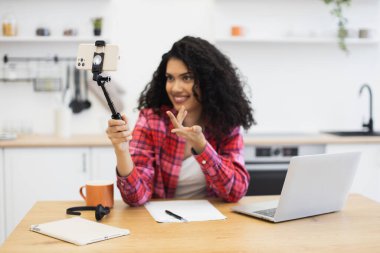 20 'li yaşlarda Afrikalı bir kadın akıllı telefon tripod dizüstü bilgisayar kullanarak mutfakta video içeriği oluşturuyor.