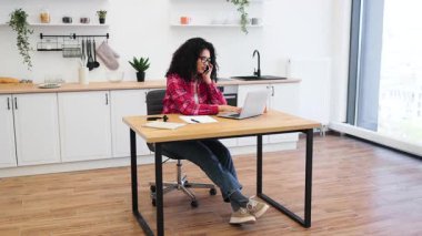 Parlak mutfakta masasında oturan Afrikalı kadın teknolojiyi kullanıyor, odaklanmış ve profesyonel görünüyor. Sahne uzaktan çalışma, üretkenlik ve konforlu ev ofisini temsil ediyor.