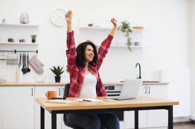 Modern mutfakta masa başında oturan gülümseyen genç Afrikalı kadın, el kaldırdı kutlama başarısını bilgisayardaki uzaktan çalışma sırasında, neşeli bir ifade