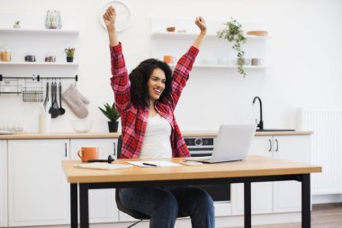Modern mutfakta masa başında oturan gülümseyen genç Afrikalı kadın, el kaldırdı kutlama başarısını bilgisayardaki uzaktan çalışma sırasında, neşeli bir ifade