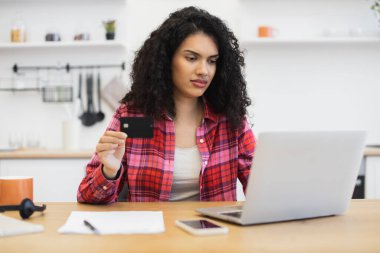 Ekose gömlekli Afrikalı genç bir kadın internette dizüstü bilgisayarla alışveriş yapıyor. El ele tutuşan kredi kartı, ifade odaklı. Çevrimiçi ticaretin modern yaşam tarzı, teknoloji kullanımı.
