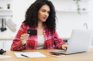 Kırmızı ekose gömlek giyen genç Afrikalı bir kadın online ödeme ve finans yönetimi için kredi kartı ve laptop kullanıyor, akıllı telefon ve belgelerle mutfaktaki ahşap masada oturuyor.
