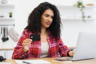 Bilgisayarı ve kredi kartını kullanarak online alışveriş yapan Afrikalı bir kadın. Kareli gömlek giyiyor, güvenli bir online işlemde, kolaylık gösteriyor..