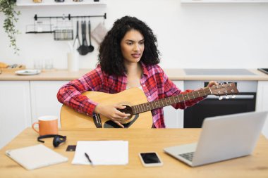 Uzun kıvırcık saçlı, gündelik kıyafetleri olan genç bir kadın mutfakta gitar çalıyor. Akustik gitar, dizüstü bilgisayar içeren teknoloji ve öğrenme kavramı, notlar.