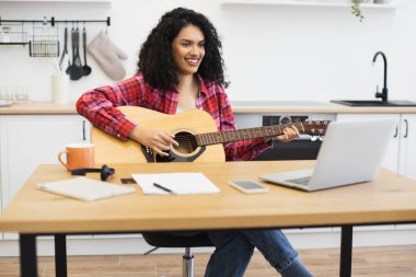 Mutfak atölyesinde sanal ders sırasında akustik gitar çalan gülümseyen genç bir kadın. Yirmili yaşlarda kadın dizüstü bilgisayarla müzik çalışmasından zevk alıyor. Yetenek ve tutku gösteriyor..