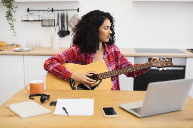 Kıvırcık saçlı, gitar çalan genç bir kadın. Mutfakta dizüstü bilgisayar başında ders izliyor. Evde öğrenim ortamı, yaratıcı kendini geliştirme, kişisel gelişimi vurgulayan kapalı alan aktivitesi.
