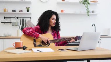Uzun kıvırcık saçlı, gündelik kıyafetleri olan genç bir kadın mutfakta gitar çalıyor. Akustik gitar, dizüstü bilgisayar içeren teknoloji ve öğrenme kavramı, notlar.