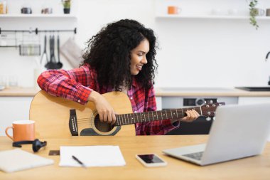 Kıvırcık saçlı, 20 'li yaşlarda, kırmızı tişörtlü, evde gitar çalan genç bir kadın.
