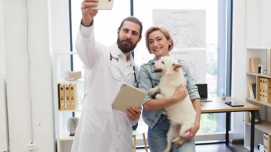 Erkek veteriner klinikte gülümserken köpek tutan kadın evcil hayvan sahibiyle selfie çekiyor.