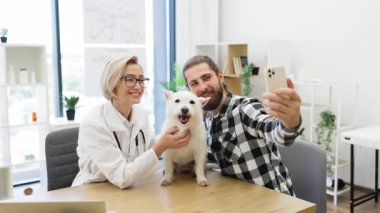 Sakallı adam gülümseyen kadın veterinerle selfie çekiyor. Küçük, tüylü, tüylü bir köpek tutuyor. Beyaz kadın 30 'lu yaşların sonlarında ve erkek evcil hayvan bakım ortamında dostça bir bağ kuruyor..