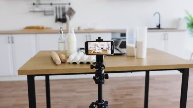 Parlak mutfak sahnesi, yemek pişirme videosunu kaydeden bir telefonu tripodun üzerinde gösteriyor. İçindekiler arasında yumurta, süt, un ve diğer malzemeler yer alıyor ve modern kamera ekipmanlarıyla mutfak içeriği oluşturulması öneriliyor..