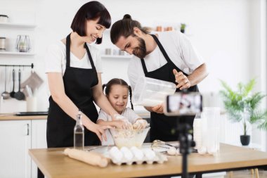 Üç kişilik beyaz bir aile mutfakta yemek pişiriyor. Siyah önlük giyip çocuklara yardım ediyorlar. Sahne, modern bir ev mutfağı ile aile bağlarını, aşçılık faaliyetlerini ve yemek bloglarını temsil ediyor.