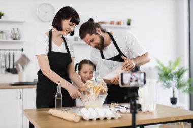 Beyaz erkek, kadın ve çocuk mutfakta siyah önlük giyerek yemek pişirme sürecini kaydediyor..