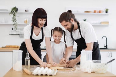 Kafkasyalı anne-baba ve kız mutfakta yemek pişiriyor, merdaneli hamuru yuvarlıyorlar.