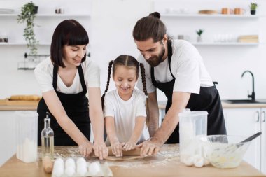 Kafkasyalı baba, anne, kız mutfakta birlikte yemek pişiriyor, hamuru neşeyle saçıyor. Parlak, modern iç mekanlarda pişirme etkinliği üzerine aile bağı..