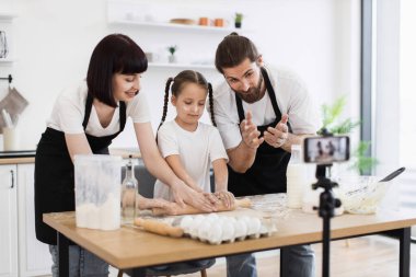 Evde yemek pişirirken hamuru yoğuran iki yetişkin ve bir çocuktan oluşan beyaz bir aile. Önlük giyiyorlar, takım çalışması ve yetenek yapıyorlar, aile bağlarını geliştirmek için..