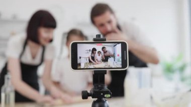 Kafkasyalı ebeveynler ve genç kız birlikte hamur yoğurarak blog yazarları olarak içerik yaratıyorlar. Çevrimiçi yemek pişirme dersi için cep telefonunu tripodda kullanarak neşeli ve çekici mutfak anları yakalıyorsun..