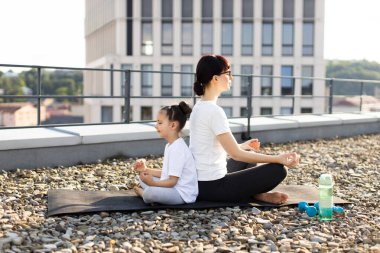 Kafkasyalı yetişkin kadın ve genç kız sırt sırta oturup çatıda meditasyon yapıyorlar. Güneşli bir günde yoga ve farkındalık yaparlar..