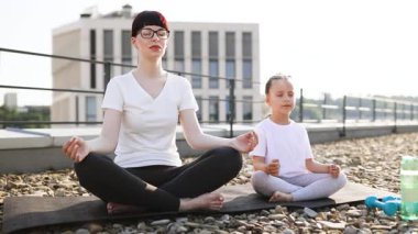 Anne ve küçük kızı sabah sükuneti içinde Lotus pozisyonunda birlikte meditasyon yapıyor, rahatlamayı, odaklanmayı ve farkındalığı teşvik ediyor. Sakinleştirici atmosferi olan huzurlu şehir merkezi.
