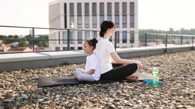 Kafkasyalı yetişkin kadın ve genç kız sırt sırta oturup çatıda meditasyon yapıyorlar. Güneşli bir günde yoga ve farkındalık yaparlar..