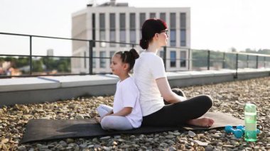 Kafkas anne ve kızı, meditasyon poziyonuyla çatıda yoga yapıyorlar. Açık havada güpegündüz sağlık ve iletişimi destekliyorlar. Sahne, fitness, sağlık, farkındalık kavramlarını vurgular
