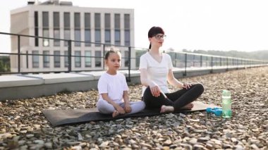 Anne ve kızı çatıda birlikte yoga yapıyorlar. Şehir sahnesinde rahatlama ve sağlıklı yaşam tarzı vurgulanıyor. İkisi de meditasyon poziyonunda bağdaş kurup aile bağlarını sergiliyor.
