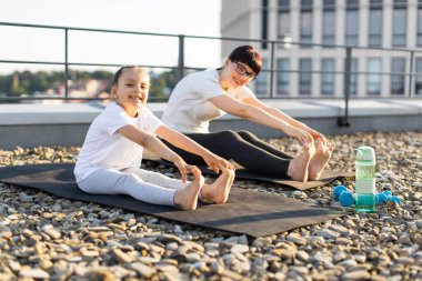 Anne ve kızı sabah güneş ışığında çatıda esneme egzersizleri yapıyorlar. Sahne fitness, sağlıklı yaşam tarzı ve aile bağlarını teşvik eder. Yoga minderlerini içerir