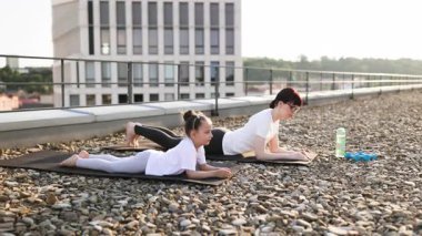 Anne ve kızı çatıda spor yapıyor. Zindelik ve bağ kurma konusunda. Parlak gün atmosferi açık hava egzersizini vurguluyor.