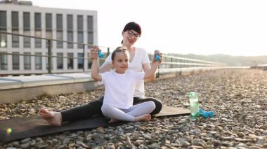 Anne ve genç kızı çatıda birlikte egzersiz yapıyor, dambıllarla spor faaliyetleri yapıyor, sağlıklı aile yaşam tarzını ve refah ve pozitif enerji için aktif alışkanlıkları destekliyor.