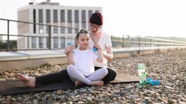 Anne kızıyla çatıda oturuyor, küçük dambıllar kullanarak spor yapıyor, bağlanma zamanının tadını çıkarıyor. Çocuk ebeveynlerinden uygun egzersiz teknikleri öğreniyor.